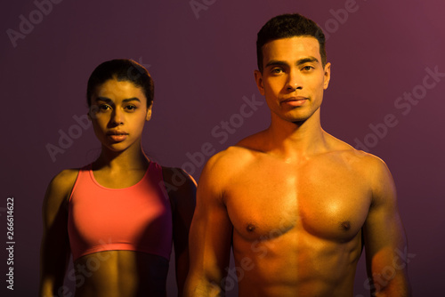 attractive african american woman in pink sports bra and shirtless mixed race man looking at camera on purple background