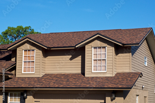 View of generic brown two story American house against blue sky with gables windows.