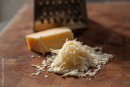 Grated italian parmesan cheese on wooden chopping board with a block of parmasan and a grater in the background. Close up photo with selective focus.