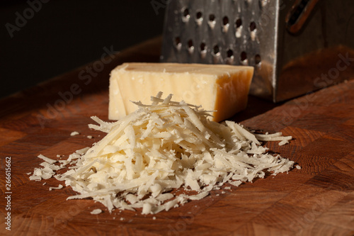 Grated italian parmesan cheese on wooden chopping board with a block of parmasan and a grater in the background. Close up photo with selective focus.