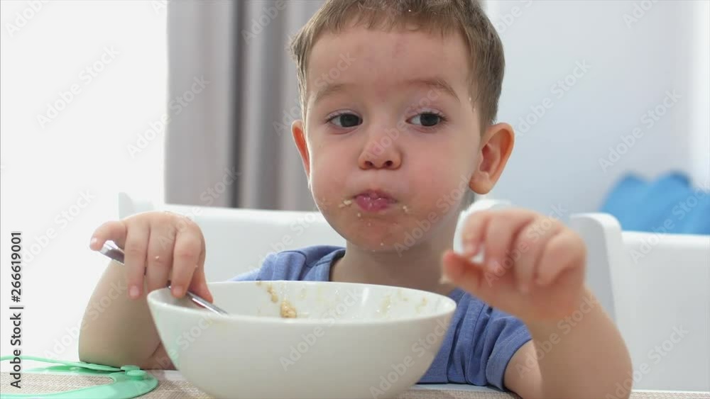Small Cute Child is Sitting at a Table and eat his own oatmeal, the baby eats willingly. Concept Happy Childhood. Small Cute Child is Sitting at a Table and eat his own oatmeal, the baby eats willingly. Concept Happy Childhood.