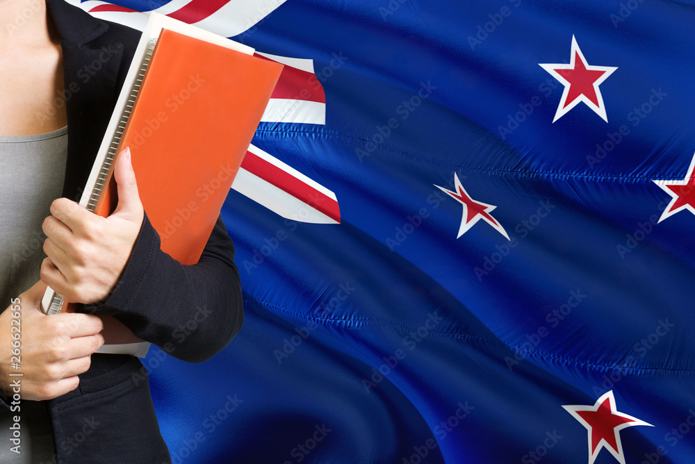 Learning language concept. Young woman standing with the New Zealand flag in the background. Teacher holding books, orange blank book cover.