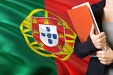 Learning Portuguese language concept. Young woman standing with the Portugal flag in the background. Teacher holding books, orange blank book cover.