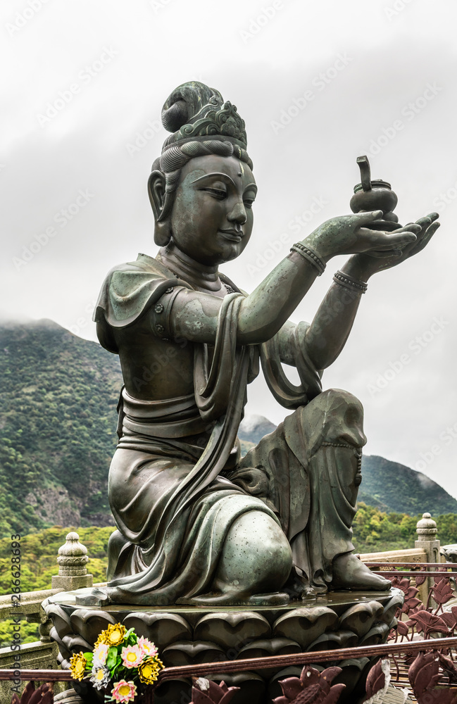 Hong Kong, China - March 7, 2019: Lantau Island. Closeup, One of the Six Devas offers ointment to Tian Tan Buddha. Bronze statue seen from front with green hill and rainy sky in back.