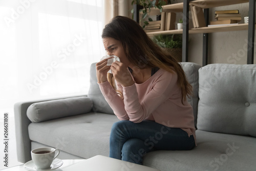 Unhealthy woman sitting on couch holding tissue wiping her nose