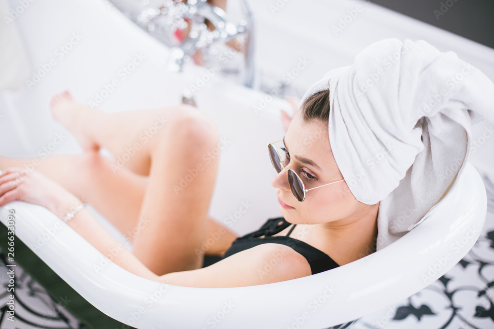 Obraz premium Young caucasian woman with towel on head and sunglasses getting spa treatment in a beauty salon, inside an interior room. Relaxing in the bathroom in linen. The concept of body care and relaxation