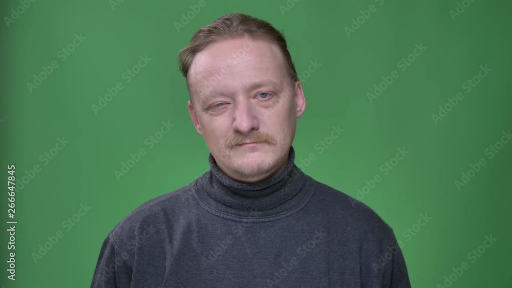 Blonde middle-aged man in gray pullover thoughtfully watching into camera and winking at it on green background. Blonde middle-aged man in gray pullover thoughtfully watching into camera and winking at it on green background.