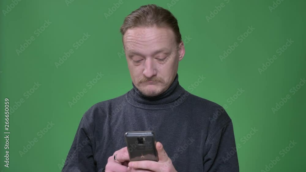 Blonde pensioner in gray pullover reacting emotionally on content in smartphone on green background. Blonde pensioner in gray pullover reacting emotionally on content in smartphone on green background.