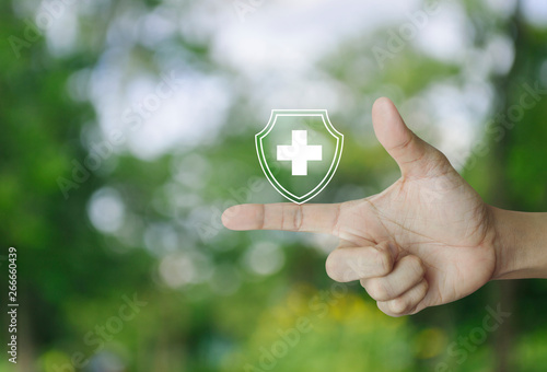 Cross shape with shield flat icon on finger over blur green tree in park, Business healthy and medical care insurance concept