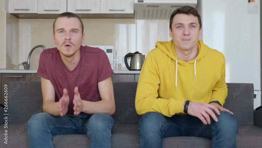 Two men sitting on the couch and watching decisive moment of football match. Young guys are in favor of sports game. The kitchen on the background. 4K footage. Two men sitting on the couch and watching decisive moment of football match. Young guys are in favor of sports game. The kitchen on the background. 4K footage.