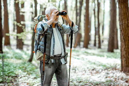 Senior man looking with binoculars while traveling with backpack in the forest