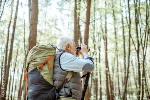 Senior man looking with binoculars while traveling with backpack in the forest