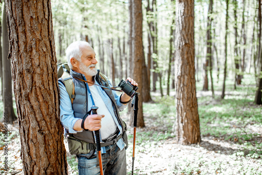Portrait of a senior man with binoculars and backpack resting near the tree while traveling in the forest Portrait of a senior man with binoculars and backpack resting near the tree while traveling in the forest