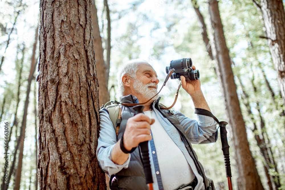 Portrait of a senior man with binoculars and backpack resting near the tree while traveling in the forest Portrait of a senior man with binoculars and backpack resting near the tree while traveling in the forest