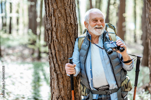 Portrait of a senior man with binoculars and backpack resting near the tree while traveling in the forest
