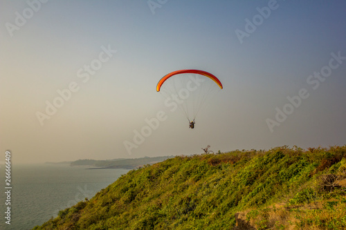 tandem paragliders on a yellow red parachute fly over the sea and green grass against a clean gray blue sky