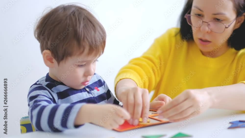 little asian boy collects puzzles with mom sitting at the desk. A cute child collects a picture from large multi-colored details. Playing with a puzzle. slow motion. Teacher playing with boy little asian boy collects puzzles with mom sitting at the desk. A cute child collects a picture from large multi-colored details. Playing with a puzzle. slow motion. Teacher playing with boy