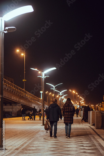 Photography Two individuals walking along a lit pathway under vibrant streetlights in an urban area