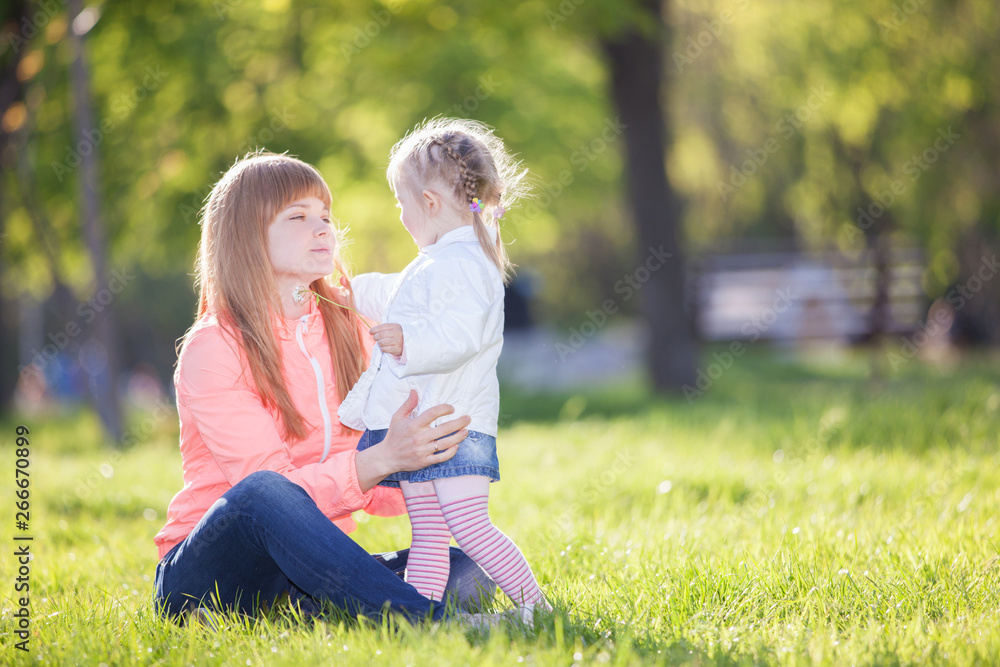 Fototapeta premium Mother and daughter in the park. Beauty nature scene with colorful background at spring season. Family outdoor lifestyle. Happy woman and cute girl relax on green grass