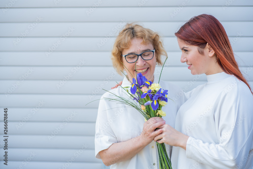 Adult daughter gives flowers to her mother outside, in the courtyard of the house. Spending time together, celebrating at home on weekends. Mothers Day. Warm intergenerational relationships Adult daughter gives flowers to her mother outside, in the courtyard of the house. Spending time together, celebrating at home on weekends. Mothers Day. Warm intergenerational relationships