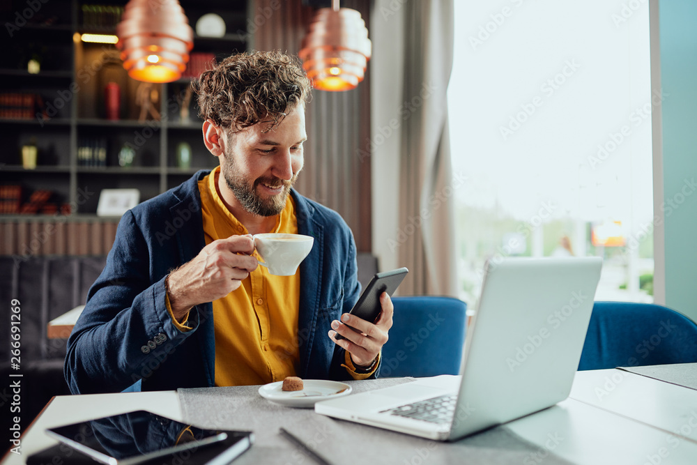 Young Caucasian bearded businessman dressed smart casual using smart phone and drinking coffee while sitting in cafe. - obrazy, fototapety, plakaty 