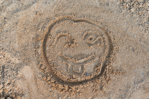 Smiley face painted on the sand with a finger. One eye winks and shows or stuck out tongue. Day, the sun is shining. Good mood, positive emotions. Rest, vacation. Friendship, good relations.
