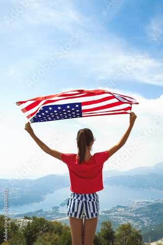 Child girl is waving American flag on top of mountain at sky background