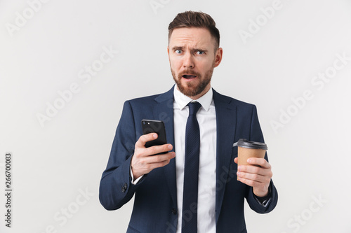 Displeased businessman posing isolated over white wall background drinking coffee using mobile phone.