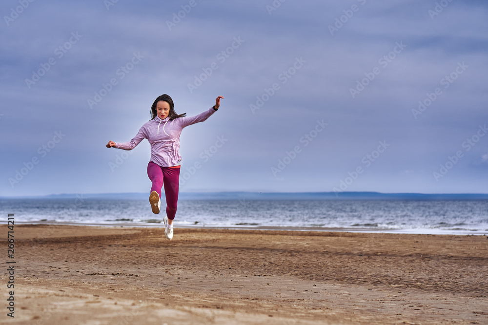 Young woman with black hair jumps while jogging. A woman is engaged in gymnastics in the spring morning on the sandy bank of a large river. Cloudy morning. Young woman with black hair jumps while jogging. A woman is engaged in gymnastics in the spring morning on the sandy bank of a large river. Cloudy morning.