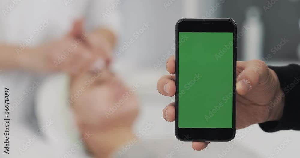 Close-up of a Man's Hand Holding Green Screen Smartphone on the osmetology Procedures Background. The oncept of Beauty, Medicine and Face Care Close-up of a Man's Hand Holding Green Screen Smartphone on the osmetology Procedures Background. The oncept of Beauty, Medicine and Face Care