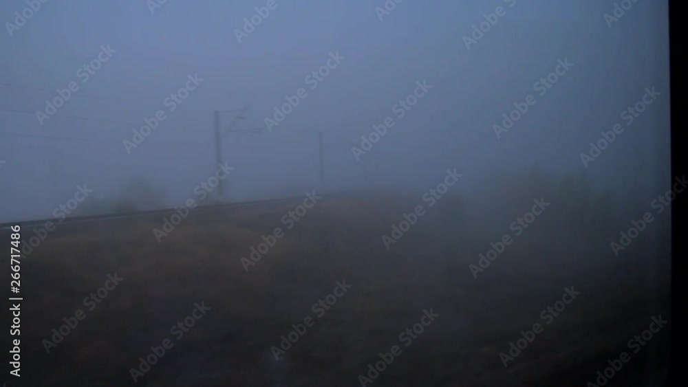 View from fast moving train passing by rural area with fields and trees covered in thick fog, and wide road at dusk