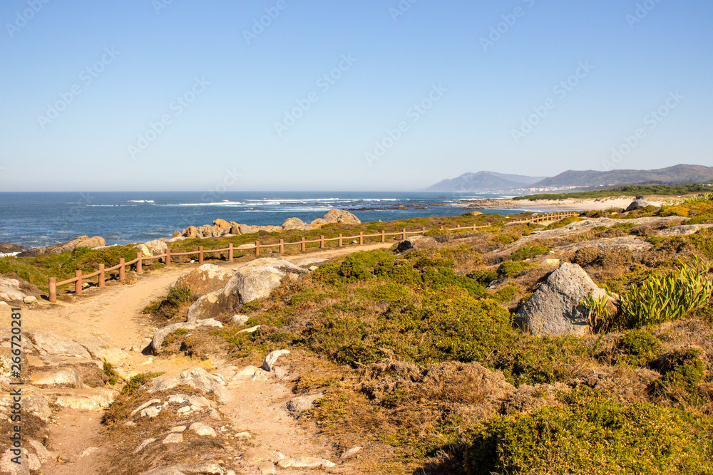 Walkway with fence along Atlantic Ocean coast with mountain on background. Portugal nature. Moss and grass on rocks at seaside. Wide beach with waves and path on Camino de Santiago. Scenic seascape.