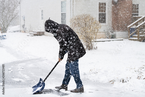 Young man in winter coat cleaning shoveling driveway street from snow in heavy snowing snowstorm with shovel by residential house and snowflakes falling