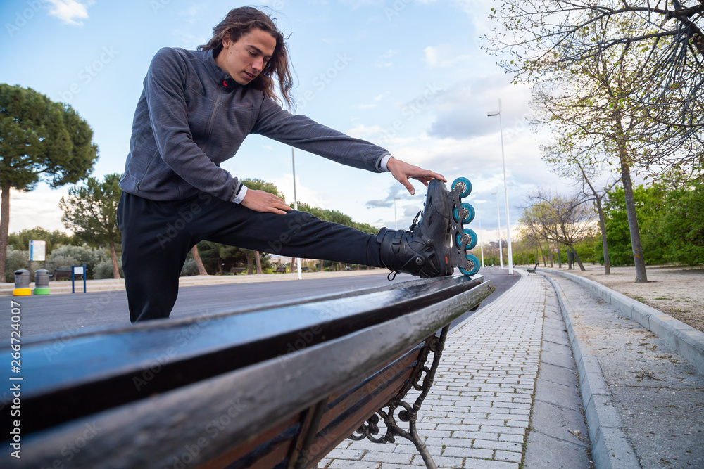 From below side view of young cool man in sportswear with roller skates stretching on bench in park From below side view of young cool man in sportswear with roller skates stretching on bench in park