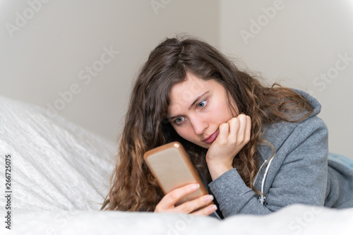 Portrait closeup of bored young woman girl lying down on bed with mobile phone on social media