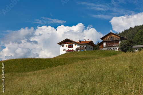 A house with flower terraces in a mountainous area against a yellow-green hilly meadow, forest, white clouds, blue sky on a bright sunny day