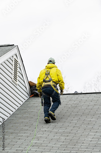 House during day with gray color home and construction man in yellow uniform walking on roof shingles and ladder during repair