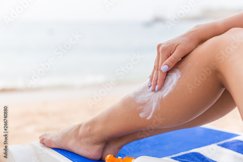 Young woman is having rest on the sunbed at the beach and protects her skin applying sunblock on her leg