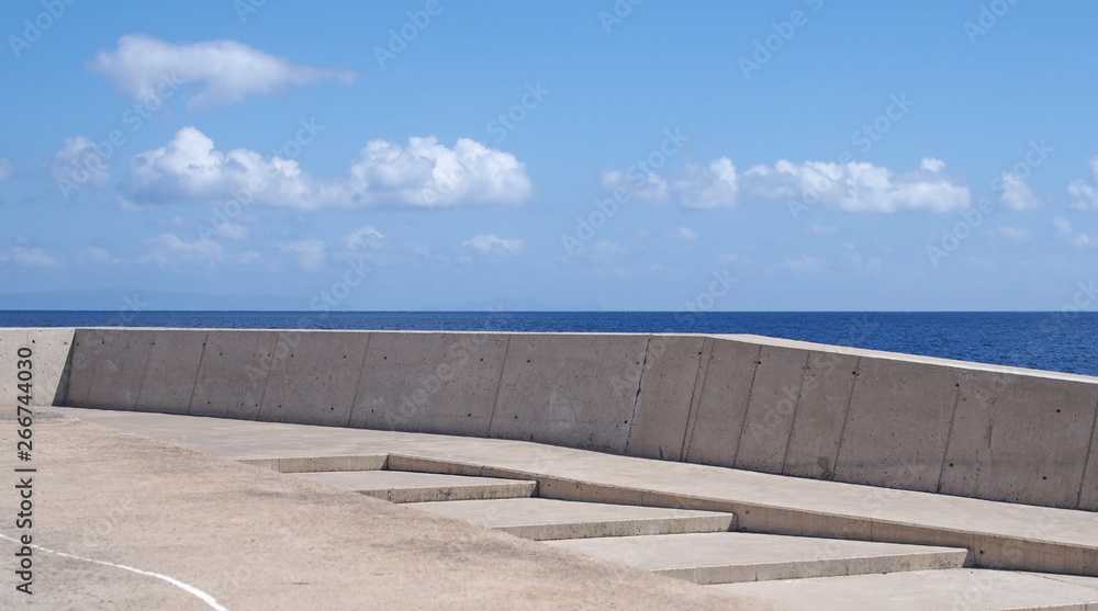 long image of a concrete sea wall with steps against a calm blue sea and sunlit blue sky