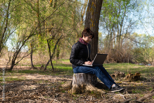 Girl using lap top among nature. Time online. Is sits on a stump near a tree