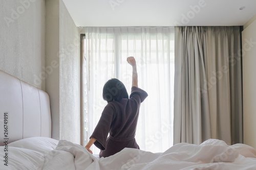 Woman stretching in bed after waking up, back view. Woman sitting near the big white window while stretching on bed after waking up with sunrise at morning, back view.