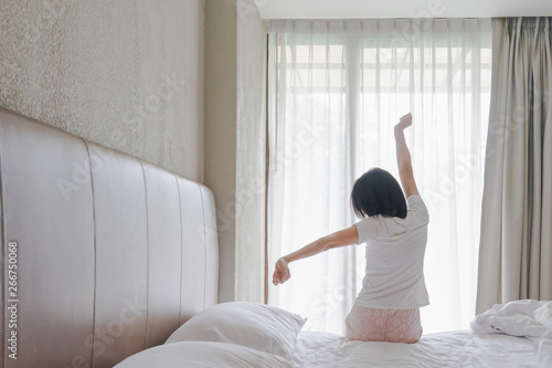 Fototapeta Naklejka Na Ścianę i Meble -  Woman stretching in bed after waking up, back view. Woman sitting near the big white window while stretching on bed after waking up with sunrise at morning, back view.