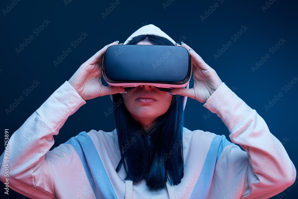 brunette girl in hood touching virtual reality headset on blue brunette girl in hood touching virtual reality headset on blue