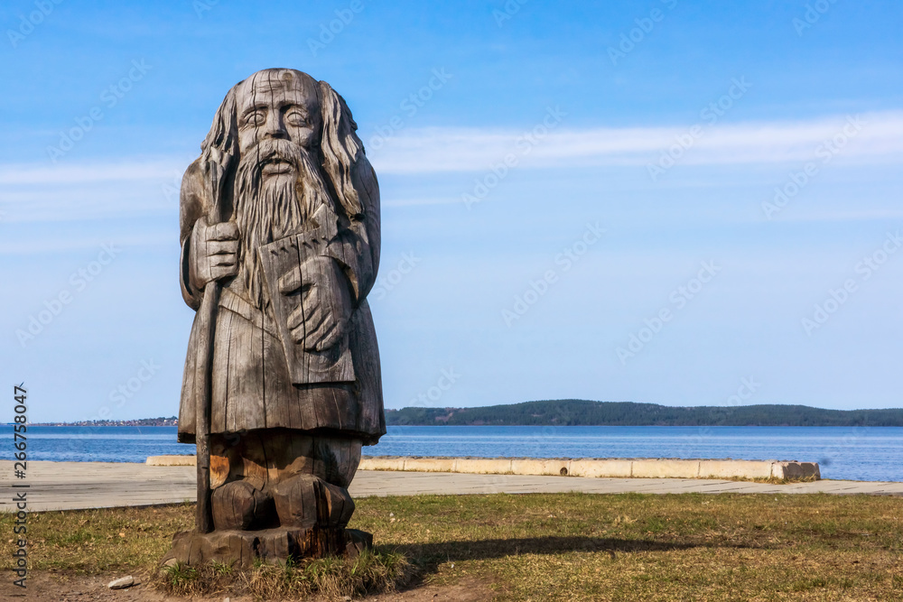 Wooden sculpture of old man in bast shoes with gusli and staff on lake shore