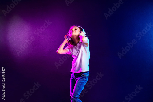 Girl listening to music in headphones on dark colorful background. Cute child enjoying happy dance music, close eye's and smile posing on studio background wall.