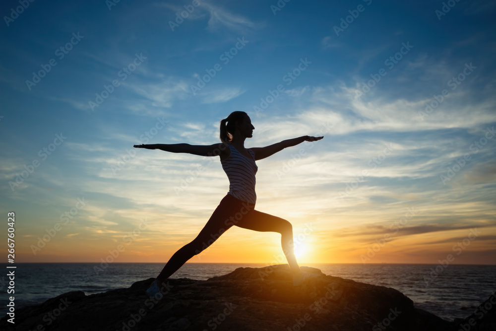 Silhouette of young woman practicing yoga standing in warrior pose on the beach at sunset.
