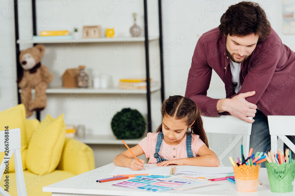 Fototapeta premium handsome father standing near cute daughter drawing at home