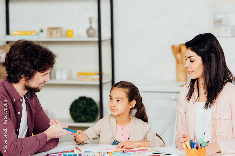 Fototapeta premium happy father giving felt pen to cute daughter near attractive wife