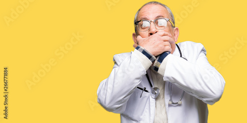 Handsome senior doctor man wearing medical coat shocked covering mouth with hands for mistake. Secret concept.