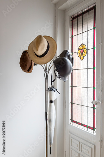 Perchero metálico sobre el que están colgados sombrero Panamá, cascos bici, gorro y paraguas sobre un fondo de ventana con vidriera photo